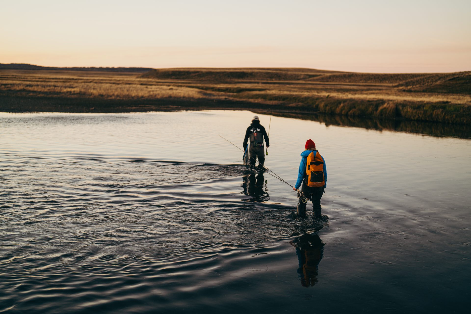 Two anglers wading across a shallow river, one is wearing a yellow Loop 25L dry backpack